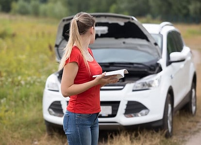 A women looking at her broke down white car holding the manual.