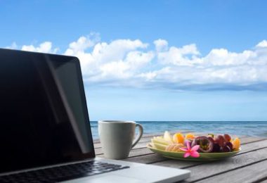 Laptop computer on an outside table with a coffee cup and fruit overlooking the ocean.