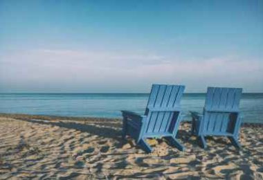 Two blue wooden chairs on a beach overlooking the ocean.